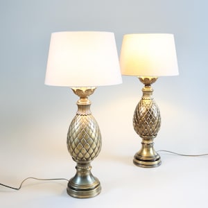 Puede incluir: Un par de lámparas de mesa doradas con pantallas blancas. Las bases de las lámparas tienen forma de piña.