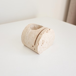 May include: A beige, semi-circular desk organizer made of stone. It features a circular hole on top and two vertical slots on the front. The organizer is placed on a white surface.