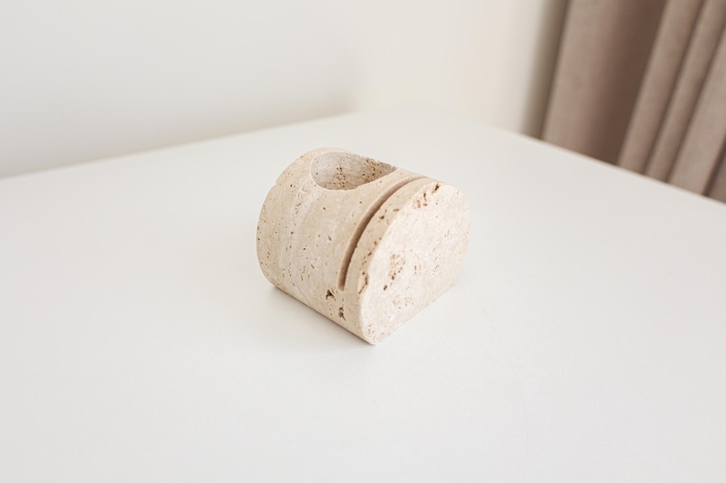 May include: A beige, semi-circular desk organizer made of stone. It features a circular hole on top and two vertical slots on the front. The organizer is placed on a white surface.