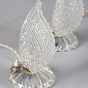 May include: A pair of clear glass table lamps featuring flame-shaped shades with a textured, cross-hatched pattern. Each lamp sits on a circular, fluted glass base and includes a light-colored twisted electrical cord with a small white switch.
