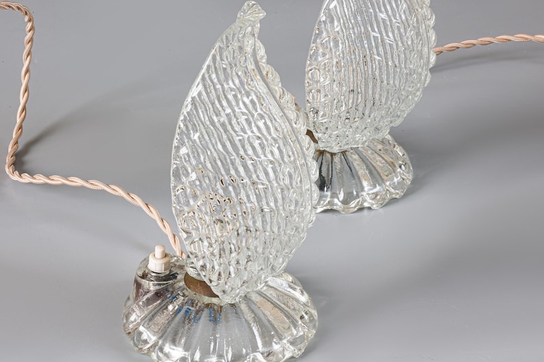 May include: A pair of clear glass table lamps featuring flame-shaped shades with a textured, cross-hatched pattern. Each lamp sits on a circular, fluted glass base and includes a light-colored twisted electrical cord with a small white switch.