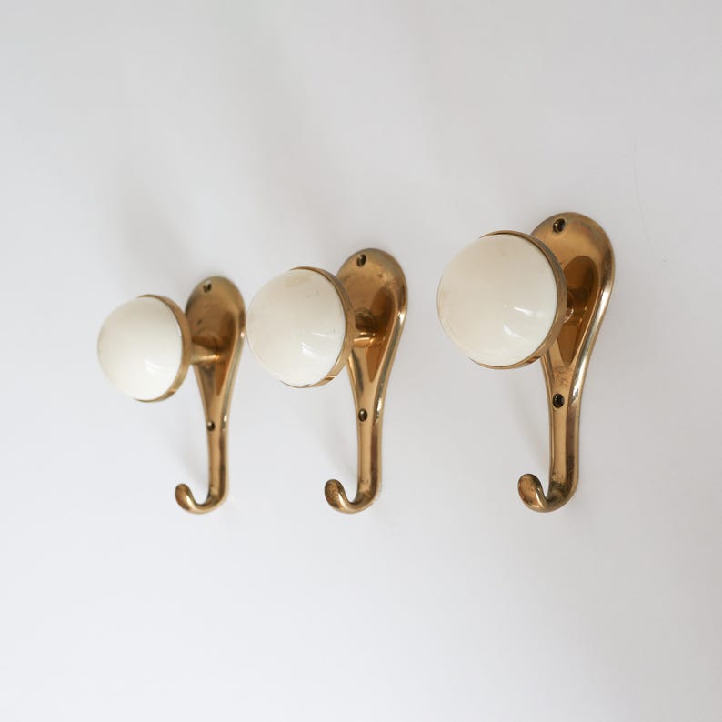 May include: Three vintage brass wall hooks with white ceramic knobs. The hooks have a curved design and are mounted on a white wall. The hooks are a classic design, suitable for coats, hats, or other items.