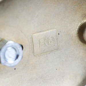 May include: Close-up of a brass-coloured metal object with a visible nut and bolt. The object has a rectangular marking with an etched design. The metal has a slightly aged appearance, with some wear and tear visible on the surface.