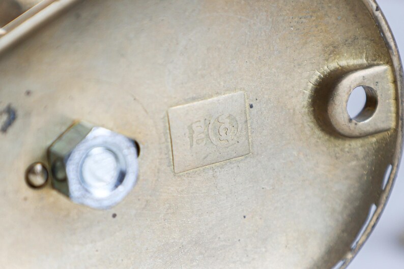 May include: Close-up of a brass-coloured metal object with a visible nut and bolt. The object has a rectangular marking with an etched design. The metal has a slightly aged appearance, with some wear and tear visible on the surface.
