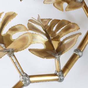 Op de afbeelding: Een gouden metalen wandsculptuur met een gestileerd ontwerp van bladeren en takken. De sculptuur is gemaakt van meerdere metalen onderdelen die aan elkaar zijn gelast om een driedimensionaal effect te cre&euml;ren.