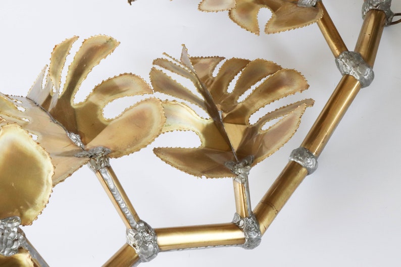 Op de afbeelding: Een gouden metalen wandsculptuur met een gestileerd ontwerp van bladeren en takken. De sculptuur is gemaakt van meerdere metalen onderdelen die aan elkaar zijn gelast om een driedimensionaal effect te cre&euml;ren.