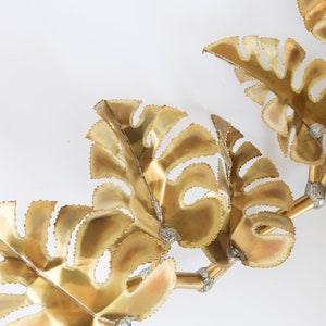Op de afbeelding: Gouden metalen wandkunst met een cluster van gestileerde bladeren. De bladeren zijn verbonden door dunne metalen stelen en hebben een getextureerd, licht verhoogd oppervlak.