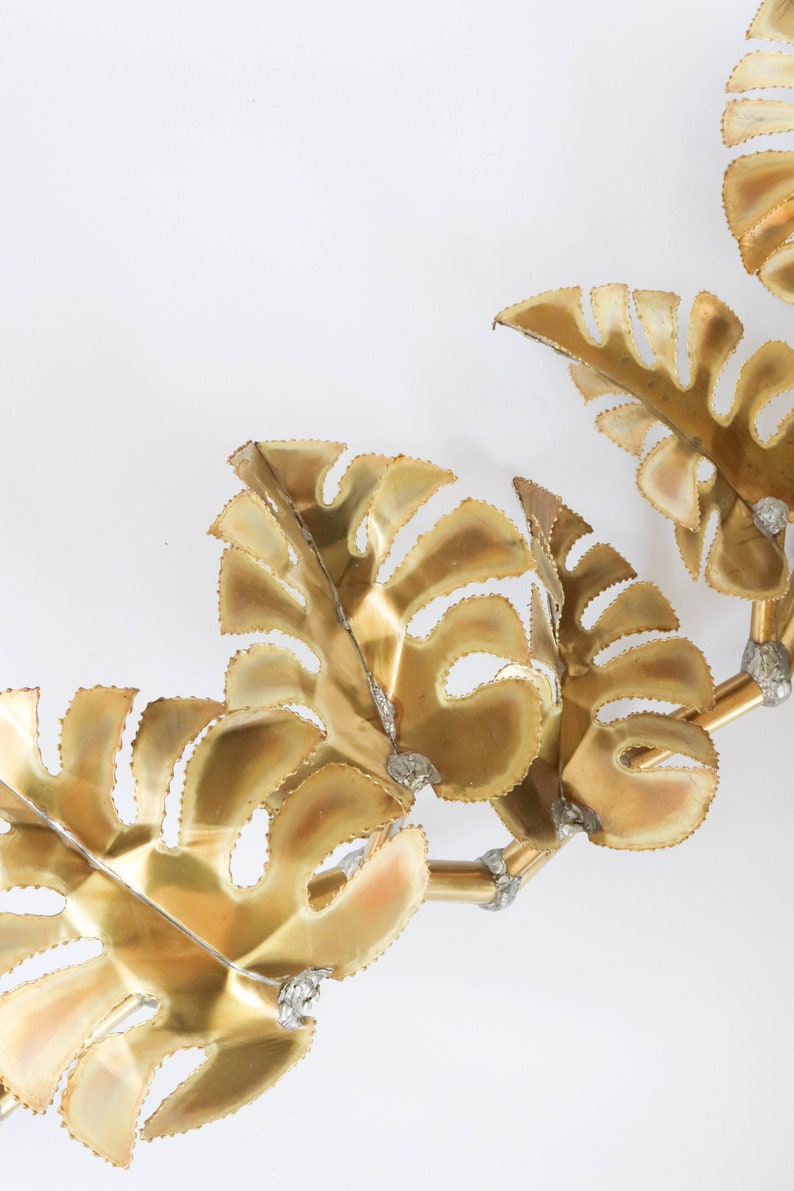 Op de afbeelding: Gouden metalen wandkunst met een cluster van gestileerde bladeren. De bladeren zijn verbonden door dunne metalen stelen en hebben een getextureerd, licht verhoogd oppervlak.