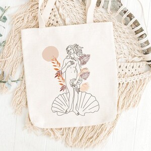 Peut inclure: Un sac fourre-tout en toile blanche avec un dessin au trait de la Naissance de V&eacute;nus, un c&eacute;l&egrave;bre tableau de la Renaissance. Le dessin est en noir et blanc avec des accents bruns et orange. Le sac fourre-tout a une sensation de coton naturel.
