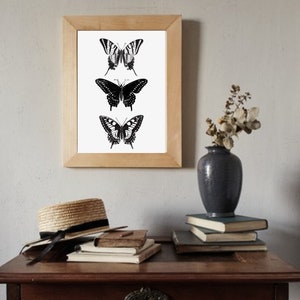 May include: A framed print of three black and white butterfly illustrations. The butterflies are arranged vertically, with the largest butterfly at the bottom and the smallest butterfly at the top. The print is hanging on a white wall above a wooden table with books and a hat.