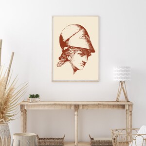 May include: A print of a woman's head wearing a helmet with a single eye in the center. The print is in shades of brown and is framed in a light wood frame. The print is hanging on a white wall above a wooden console table.