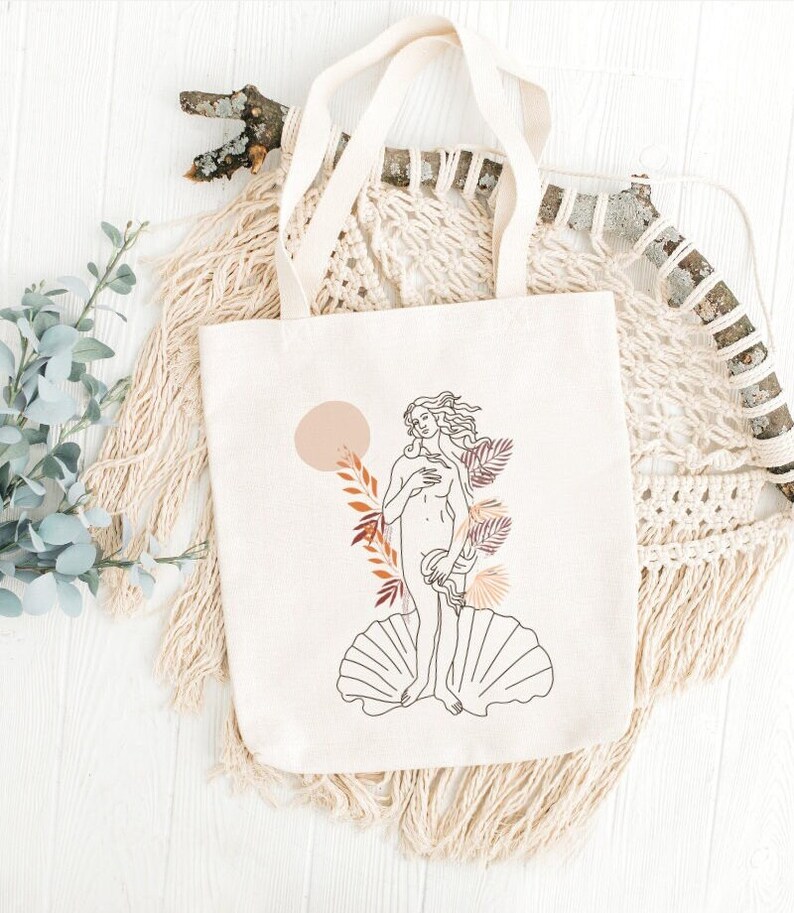 Peut inclure: Un sac fourre-tout en toile blanche avec un dessin au trait de la Naissance de V&eacute;nus, un c&eacute;l&egrave;bre tableau de la Renaissance. Le dessin est en noir et blanc avec des accents bruns et orange. Le sac fourre-tout a une sensation de coton naturel.