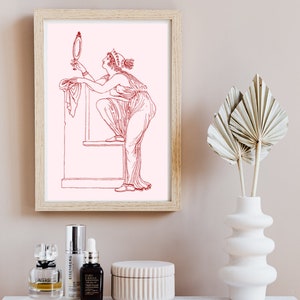 May include: A framed print of a line drawing of a woman in a long dress looking at a mirror. The drawing is in red on a pink background. The print is in a light wood frame and is hanging on a light beige wall.
