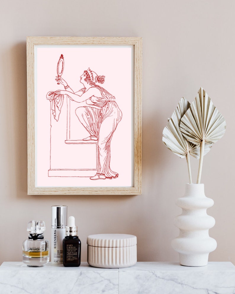 May include: A framed print of a line drawing of a woman in a long dress looking at a mirror. The drawing is in red on a pink background. The print is in a light wood frame and is hanging on a light beige wall.