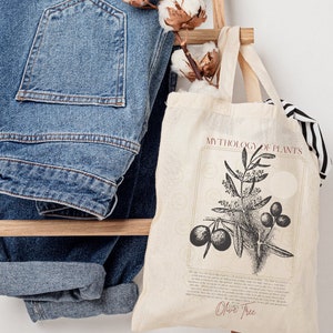 Pode incluir: Uma bolsa tote de lona branca com ilustra&ccedil;&otilde;es em preto e branco de uma oliveira e o texto "MYTHOLOGY OF PLANTS" e "Olive Tree". A bolsa tem uma al&ccedil;a marrom e est&aacute; sentada em uma escada de madeira com um par de cal&ccedil;as jeans azuis e bolas de algod&atilde;o.