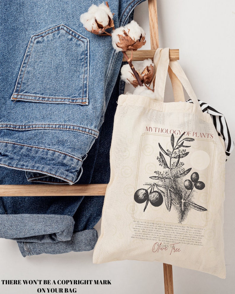 Pode incluir: Uma bolsa tote de lona branca com ilustra&ccedil;&otilde;es em preto e branco de uma oliveira e o texto "MYTHOLOGY OF PLANTS" e "Olive Tree". A bolsa tem uma al&ccedil;a marrom e est&aacute; sentada em uma escada de madeira com um par de cal&ccedil;as jeans azuis e bolas de algod&atilde;o.