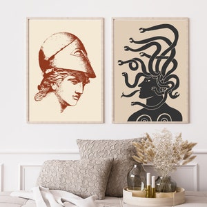 May include: Two framed prints with a minimalist design. The print on the left features a woman's head in profile wearing a helmet with a single eye. The print on the right features a woman's head in profile with snakes for hair. Both prints are in shades of brown and black on a cream background.