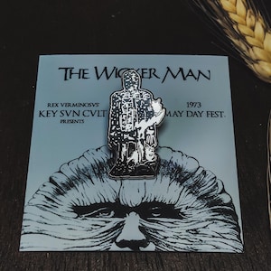 Può includere: Spilla in smalto bianco e nero raffigurante un personaggio del film "The Wicker Man", appoggiata su una carta azzurra. La carta mostra il titolo e i dettagli del film, con un'illustrazione stilizzata.