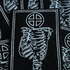Puede incluir: Parche bordado negro con un diseño de torso de esqueleto blanco y un símbolo de cruz en la parte superior. El parche rectangular tiene un borde blanco. Varios parches son visibles.