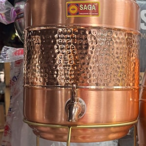 Puede incluir: Un dispensador de agua de cobre con una textura martillada en la sección media. El dispensador tiene un grifo, una tapa y se apoya en un soporte de metal dorado. El logotipo "SAGA TABLEWARE COLLECTION" es visible.