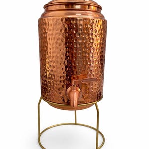 May include: A copper drinks dispenser with a hammered finish, a tap, and a lid with a gold-coloured knob. It stands on a gold-coloured metal stand. The dispenser is designed for serving drinks.
