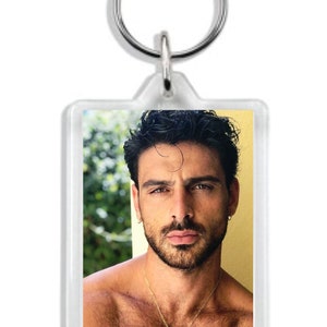 May include: A clear acrylic keychain with a photo of a man with dark hair and a beard. He is wearing a gold necklace with a pendant. The photo is set against a blurred background of green foliage. The keychain has a silver ring.