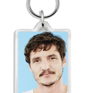 May include: A clear plastic keyring with a silver key ring. The keyring features a photo of a man with brown hair and a moustache. The man is wearing a white shirt.