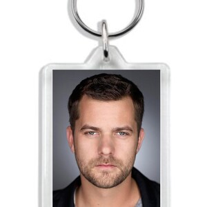 May include: A clear acrylic keychain with a photo of a man with short brown hair and a beard. He is wearing a black jacket over a gray shirt.