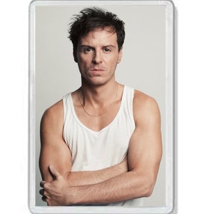 May include: A rectangular magnet featuring a man with dark hair and a serious expression. He wears a white tank top and a thin gold chain. His arms are crossed, and the background is a neutral color.