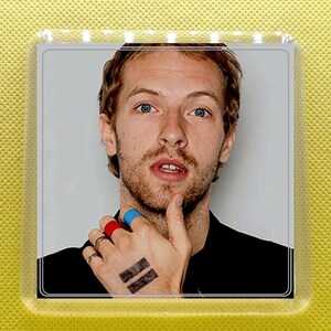 May include: A close-up portrait of a man with short brown hair and a beard. He is wearing a black shirt and has a blue, red, and yellow band on his finger. He has a tattoo on his hand that says "= =" in black ink.