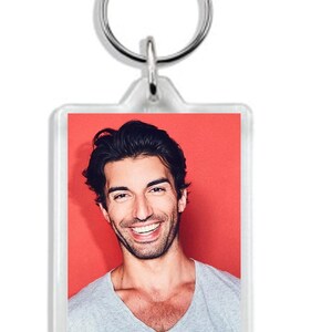 May include: A clear acrylic keychain featuring a photo of a man smiling. The man has dark hair and is wearing a grey t-shirt. The background is red.