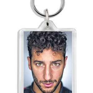 May include: A clear acrylic keyring featuring a photo of a man with curly brown hair and brown eyes. He is wearing a racing suit with a red bull logo.
