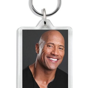 May include: A clear acrylic keychain with a photo of Dwayne Johnson smiling. The photo is in a rectangular frame with rounded corners.