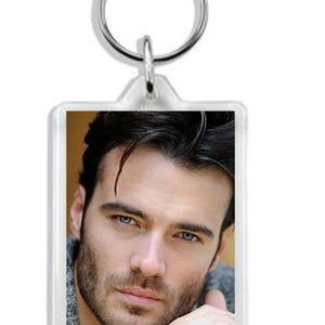 May include: Clear rectangular keychain featuring a portrait of a man with dark hair, blue eyes, and a beard. The keychain has a silver-colored metal ring at the top.