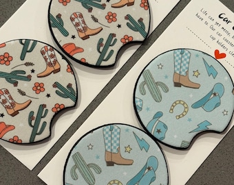 May include: Set of four car coasters with a southwestern theme. Two coasters feature a light blue background with a pattern of cowboy boots, hats, horseshoes, and stars. The other two coasters feature a light gray background with a pattern of cowboy boots, cacti, and flowers. The text "Car Coaster" is visible on the card.