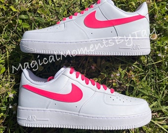 May include: White leather low-top sneakers with a bright pink Nike swoosh and laces. The shoes have "AIR" printed on the side.