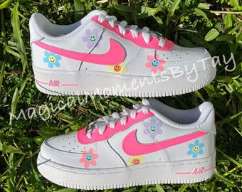May include: White low-top sneakers with a pink swoosh and a smiley face flower design. The text "AIR" is on the side of the shoe.