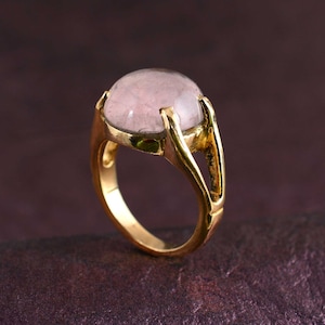 Op de afbeelding: Een gouden ring met een grote, ronde, lichtroze edelsteen. De ring heeft een uniek, open ontwerp met een gespleten band.
