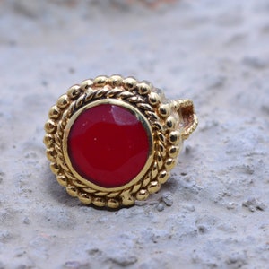 May include: A gold ring with a red gemstone set in a circular bezel. The ring has a decorative design with a rope-like pattern around the bezel.