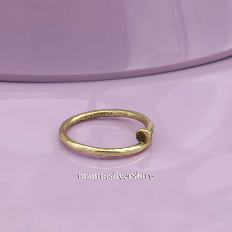 May include: A simple gold-toned ring with a nail-shaped design. The ring is on a light purple surface.