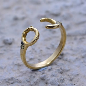 May include: A gold ring with a unique design featuring a circle and a curved shape. The ring is made of metal and has a smooth finish.