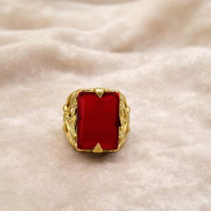 May include: A gold-colored ring with a large, rectangular, red gemstone. The ring has an ornate, woven-like design around the gemstone. The ring is set against a soft, beige background.