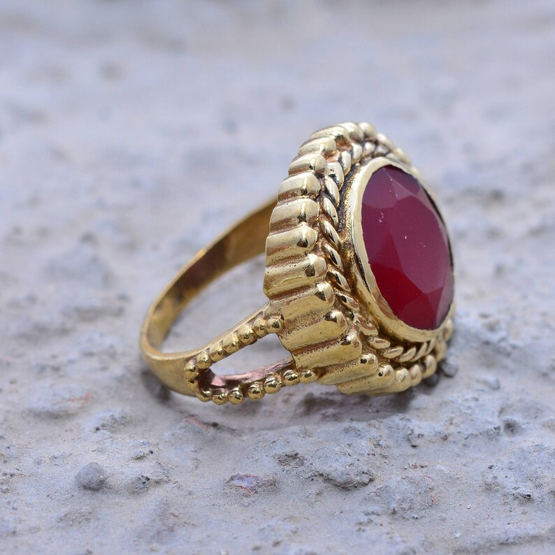 May include: A gold ring featuring a red gemstone set within an ornate, decorative setting. The ring has a substantial band and intricate detailing.