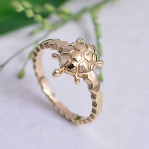Può includere: Un anello d'oro con un designo di tartaruga dettagliato. La fascia dell'anello è testurizzata con un motivo ripetitivo.