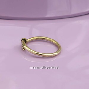 May include: A simple gold band ring with a small, textured design on the side. The ring is on a purple surface.