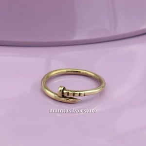 May include: A gold-toned ring with a nail design. The nail is bent at a 90-degree angle and has a textured surface.