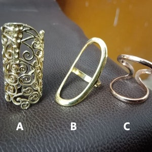 Könnte beinhalten: Drei goldfarbene Ringe. Der erste Ring ist ein breiter Band mit einem zarten, filigranen Design. Der zweite Ring ist eine einfache ovale Form. Der dritte Ring ist ein Doppelband mit einer gehämmerten Oberfläche.