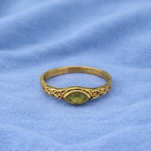 Pode incluir: Um anel dourado com uma pedra preciosa oval verde claro. O anel tem um design de filigrana ornamental na banda e ao redor da pedra preciosa. O anel está sobre um fundo de tecido azul. Joalharia.