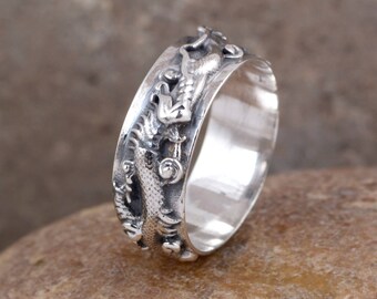 Può includere: Un anello d'argento con un designo di drago dettagliato. L'anello è largo e ha una finitura liscia e lucida.