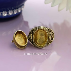 May include: A gold-colored ring with an oval setting and ornate detailing. The ring is open, with a separate, matching lid. The ring is on a purple surface. The text "mamtasilverstore" is visible.
